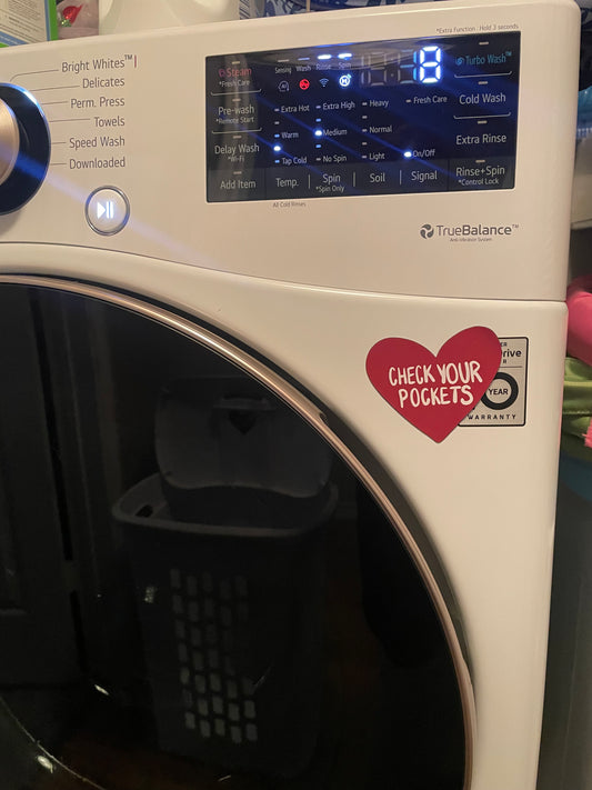 Red Heart Shaped Magnet reads “Check Your Pockets” it is displayed on a front loading washing machine